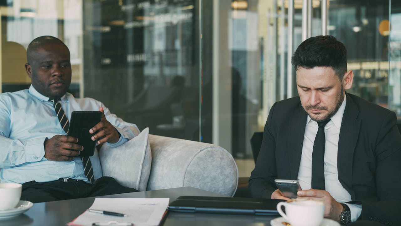 Two businessmen focused on tablets and phones in modern office lounge.