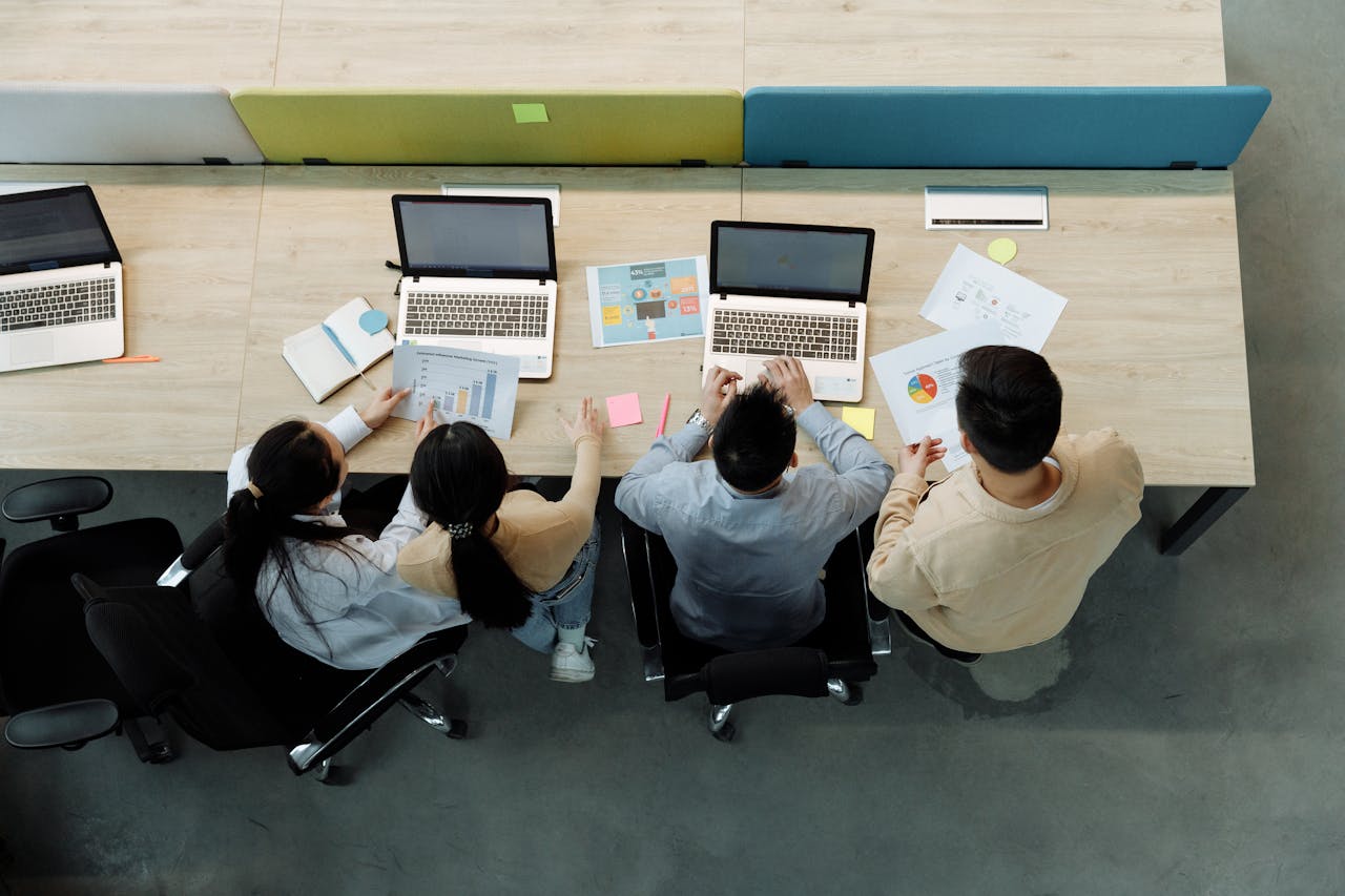 Home Aerial view of four colleagues working with laptops and documents at a shared desk.