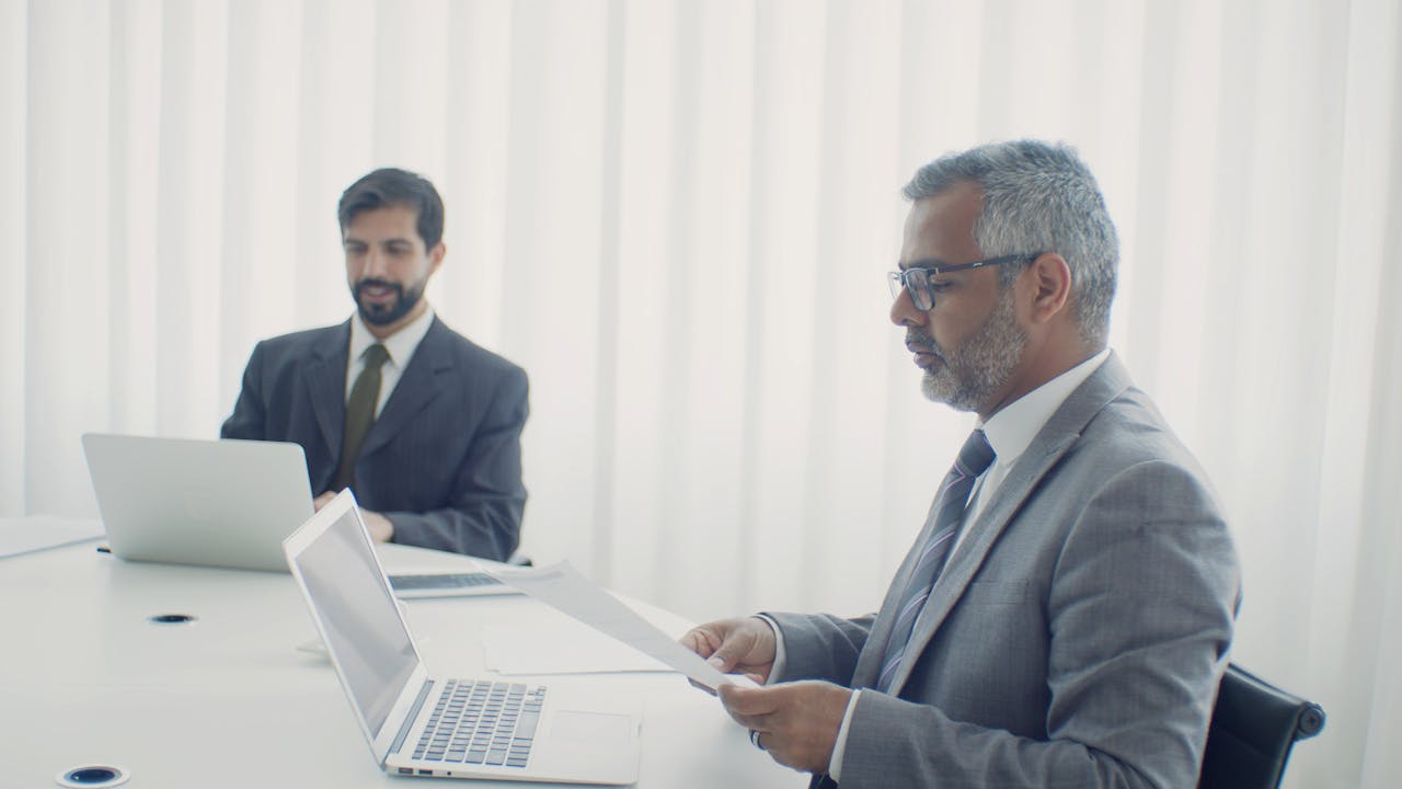 Two colleagues in a corporate setting, working on laptops and documents.